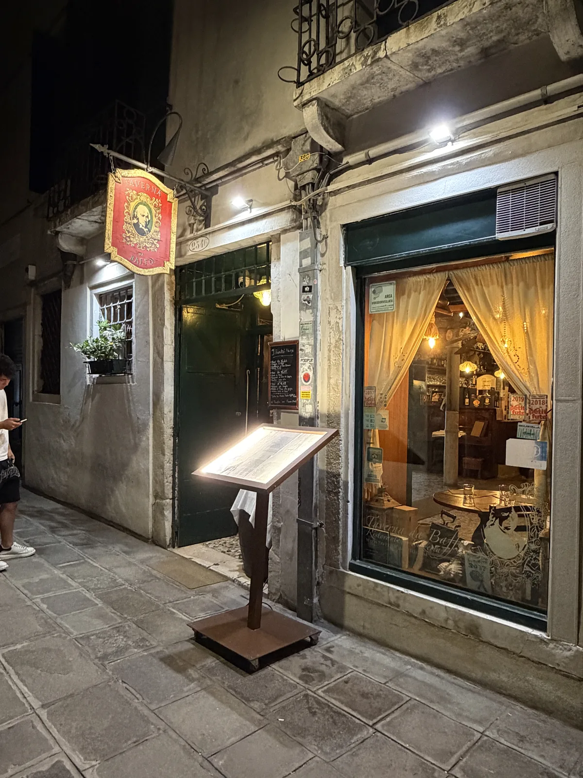 L'ingresso della Taverna da Baffo di notte, con l'insegna rossa, il leggio del menu e la vetrina illuminata